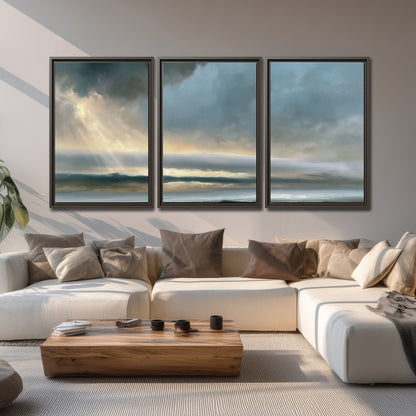 Dramatic blue-gray storm clouds breaking over a pale strip of ocean light 3 piece framed canvas wall art displayed in a living room