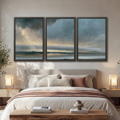 Dramatic blue-gray storm clouds breaking over a pale strip of ocean light 3 piece framed canvas wall art displayed in a bedroom