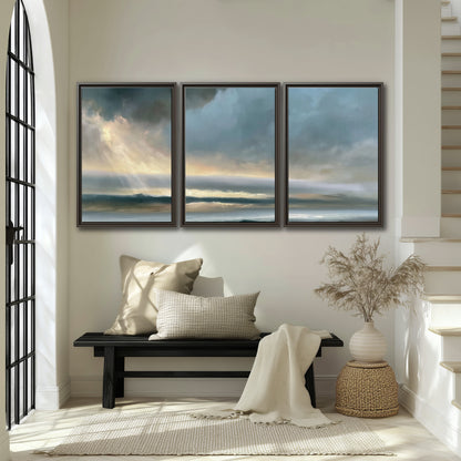 Dramatic blue-gray storm clouds breaking over a pale strip of ocean light 3 piece framed canvas wall art displayed in a entryway
