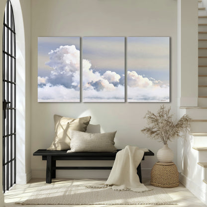 Large white clouds rising through a pale blue sky with gentle warm highlights 3 piece canvas wall art displayed in a entryway