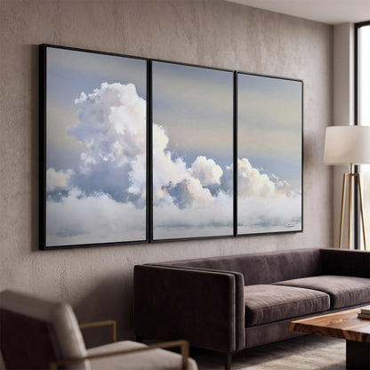 Large white clouds rising through a pale blue sky with gentle warm highlights 3 piece framed canvas wall art displayed in a modern living room