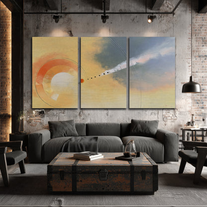 A warm orange circular form and diagonal pale streaks across a muted yellow and blue field 3 piece canvas wall art displayed in a industrial room