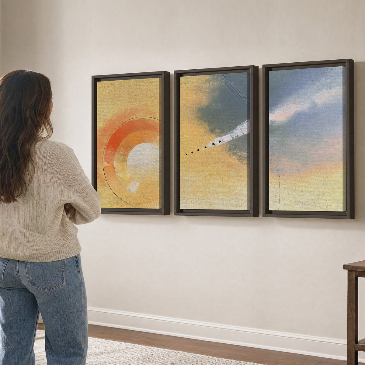 A warm orange circular form and diagonal pale streaks across a muted yellow and blue field 3 piece framed canvas wall art three panel arrangement on a wall