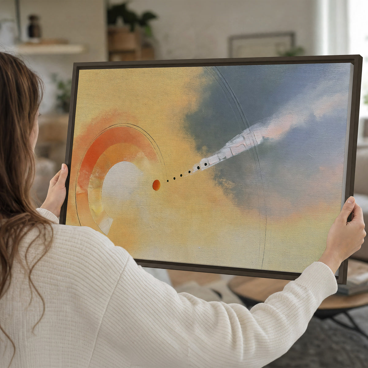 A warm orange circular form and diagonal pale streaks across a muted yellow and blue field single framed canvas wall art held in hands