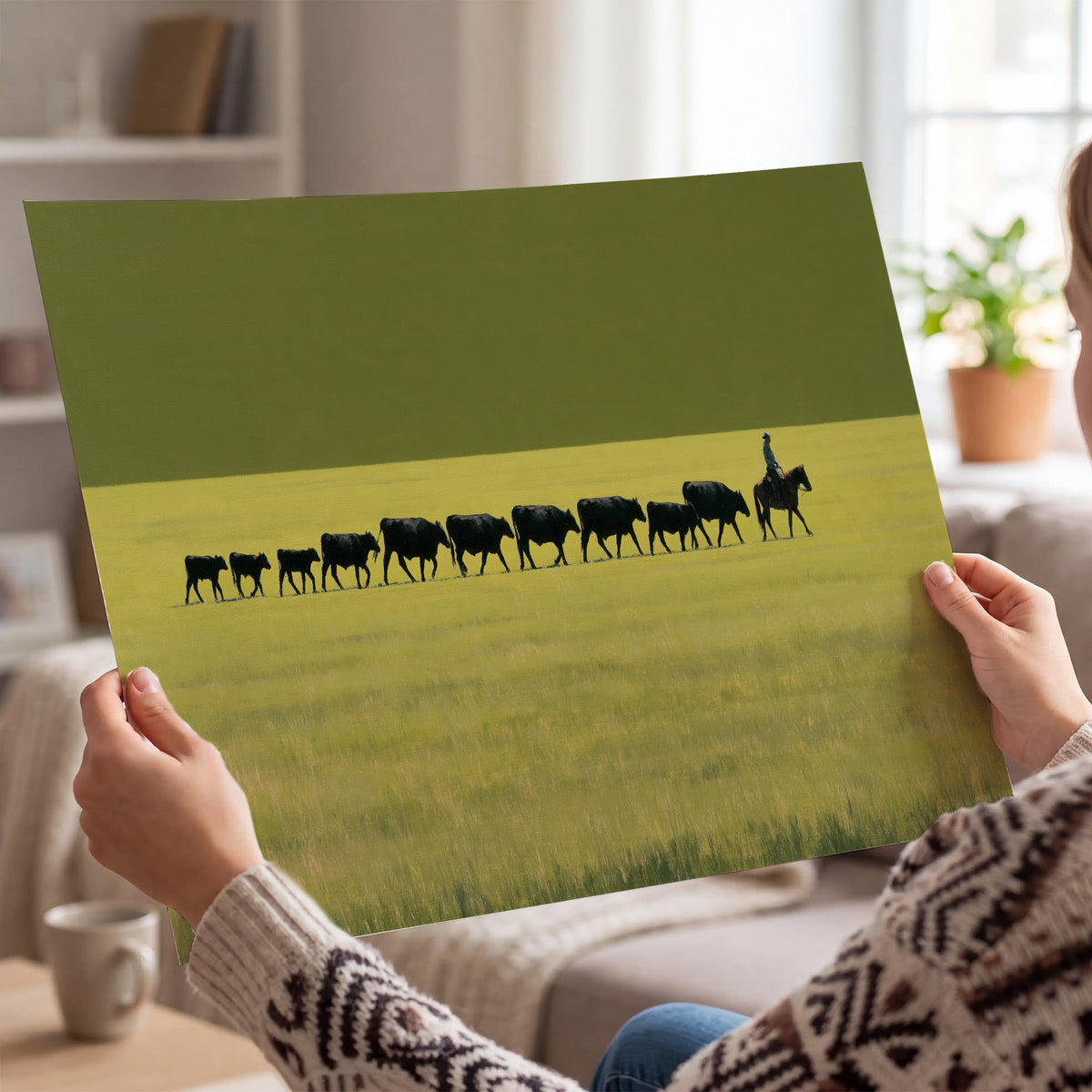 A rider leading a long line of cattle across a wide green and golden plain single poster print held in hands