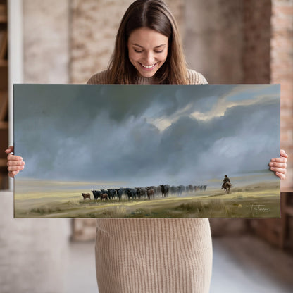 A rider and cattle moving across a sunlit grassland beneath a dramatic blue-gray sky single canvas wall art held in hands