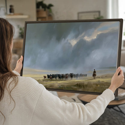 A rider and cattle moving across a sunlit grassland beneath a dramatic blue-gray sky single framed canvas wall art held in hands
