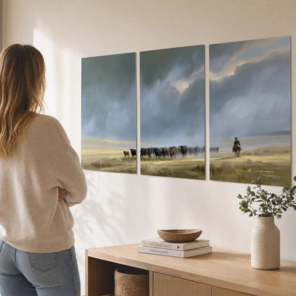 A rider and cattle moving across a sunlit grassland beneath a dramatic blue-gray sky set of 3 poster prints three panel arrangement on a wall